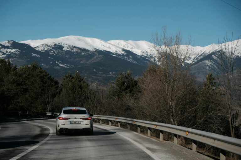 A white car driving on a curvy mountain road with snow-capped peaks in the background.